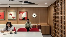 two people are sitting at a table in an office with red chairs and artwork on the walls