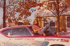 a woman sitting on the hood of a car in front of a house and tree