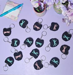 twelve black and pink dragonfly keychains are on a table next to purple flowers