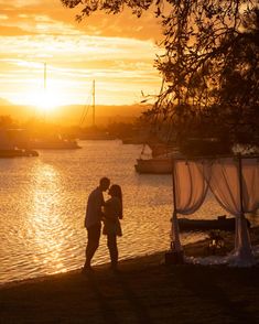 two people standing next to each other in front of a body of water at sunset