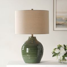 a green vase sitting on top of a white table next to a lamp and flowers