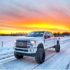 a white truck driving down a snow covered road next to a forest at sunset in the winter