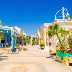 people are walking and riding bicycles on the street in front of colorful buildings with palm trees