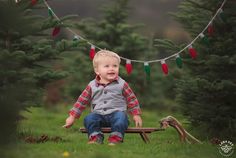 a toddler sitting on a bench in the grass with christmas lights strung above him