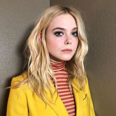 a woman with blonde hair wearing a yellow jacket and red striped turtle neck shirt, standing in front of a wall