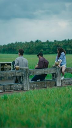 three people sitting on a wooden bench in front of a field with green grass and trees