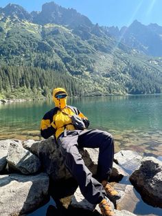 a man in yellow jacket sitting on rocks next to water with mountains in the background