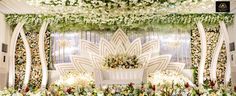 an elaborately decorated stage with white flowers and greenery on the wall, surrounded by chairs