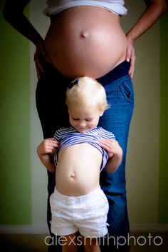 a pregnant woman standing next to a toddler who is wearing white shorts and a striped shirt