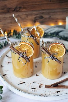 three glasses filled with orange juice and cinnamon sticks on a white plate surrounded by greenery