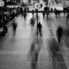 black and white photograph of people walking in an indoor area with clocks on the wall