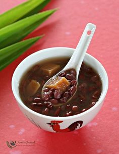 a white bowl filled with beans and meat on top of a red tablecloth next to green leaves