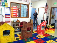 a man standing in front of a classroom filled with cardboard houses and other items on the floor