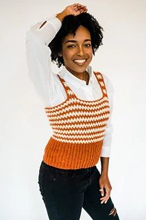 a woman in an orange and white knitted sweater smiles while standing with her hands on her head