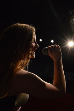 a woman singing into a microphone in front of spotlights on a dark stage with red carpet