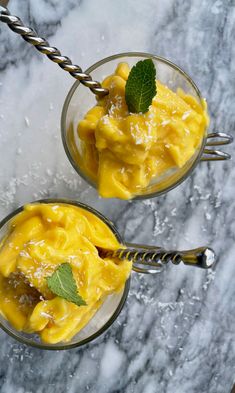 two glasses filled with yellow dessert on top of a table next to silverware and green leaves