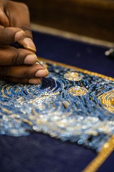 a person is working on an art project with gold and blue beads, while another hand holds a pen