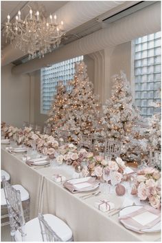 a table set up with white and pink flowers, napkins and place settings in front of christmas trees