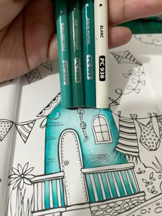 a hand holding two markers in front of a coloring book with an image of a house on it