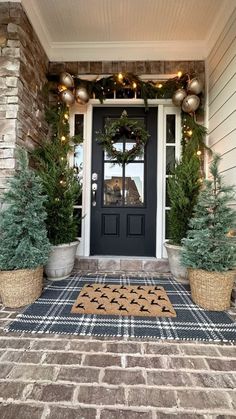 two potted trees are on the front porch with christmas lights hanging above them and a door mat