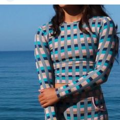 a woman standing next to the ocean wearing a blue and white checkered top with her arms crossed