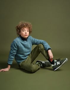 a young boy sitting on the ground wearing green pants and a blue sweater with his hands in his pockets