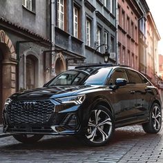 a black suv parked on the side of a cobblestone road next to tall buildings