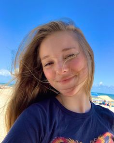 a girl with long hair standing on the beach and smiling at the camera while wearing a dragon t - shirt