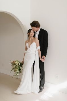 a bride and groom standing next to each other in front of a white wall holding hands