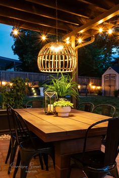 an outdoor dining table with lights hanging from it's ceiling and chairs around it