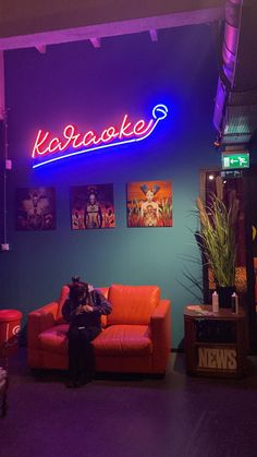 a person sitting on an orange couch in front of a neon sign that reads kalabake