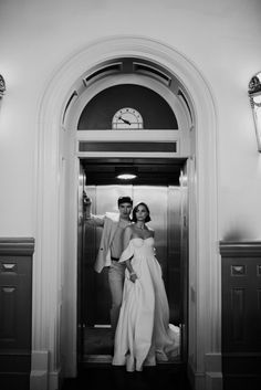 black and white photo of two people in front of an open elevator with clock on the wall