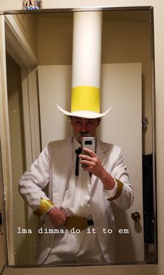 a man in a white suit and yellow hat holding a cell phone while standing in front of a mirror