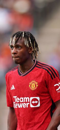 a man with dreadlocks standing in front of a soccer field wearing a red shirt