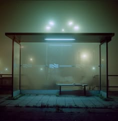 an empty bus stop at night with the lights on and fog covering the glass walls