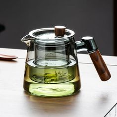 a glass tea pot with a wooden handle on a table next to a cup of green tea