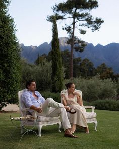 a man and woman sitting on lawn chairs in front of trees with mountains in the background