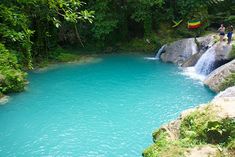 people are standing on the edge of a waterfall with blue water and green vegetation surrounding it