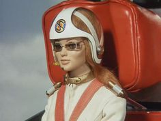 a barbie doll wearing a helmet and glasses sitting in a chair with luggage behind her