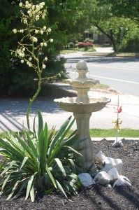 a small fountain in the middle of a flower bed next to a street and trees