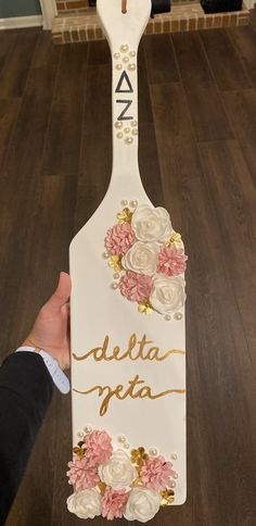 a person holding up a wooden sign with flowers on it that says delta delta in gold lettering