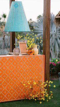 an orange table with a blue lamp on top and flowers in the vase next to it