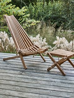 two wooden chairs sitting on top of a wooden deck next to tall grass and bushes