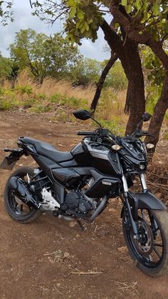 a black motorcycle parked next to a tree on a dirt road in front of some trees