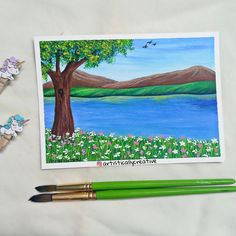 two green pencils are next to an art card with water and trees on it