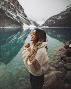 a woman standing in front of a body of water with mountains in the background and snow on the ground