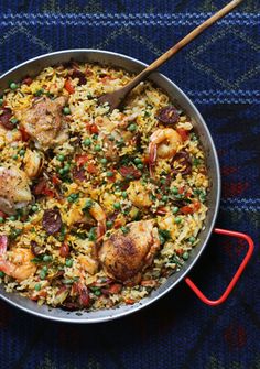 a pan filled with rice and shrimp on top of a blue table cloth next to a wooden spoon