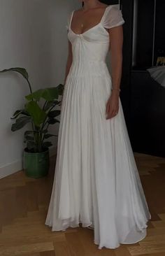 a woman in a white dress standing on a wooden floor next to a potted plant