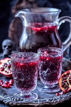 two glasses filled with red liquid next to a pitcher and pomegranates