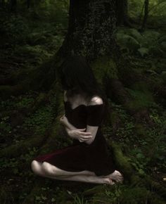 a woman sitting on the ground in front of a tree with moss growing around her